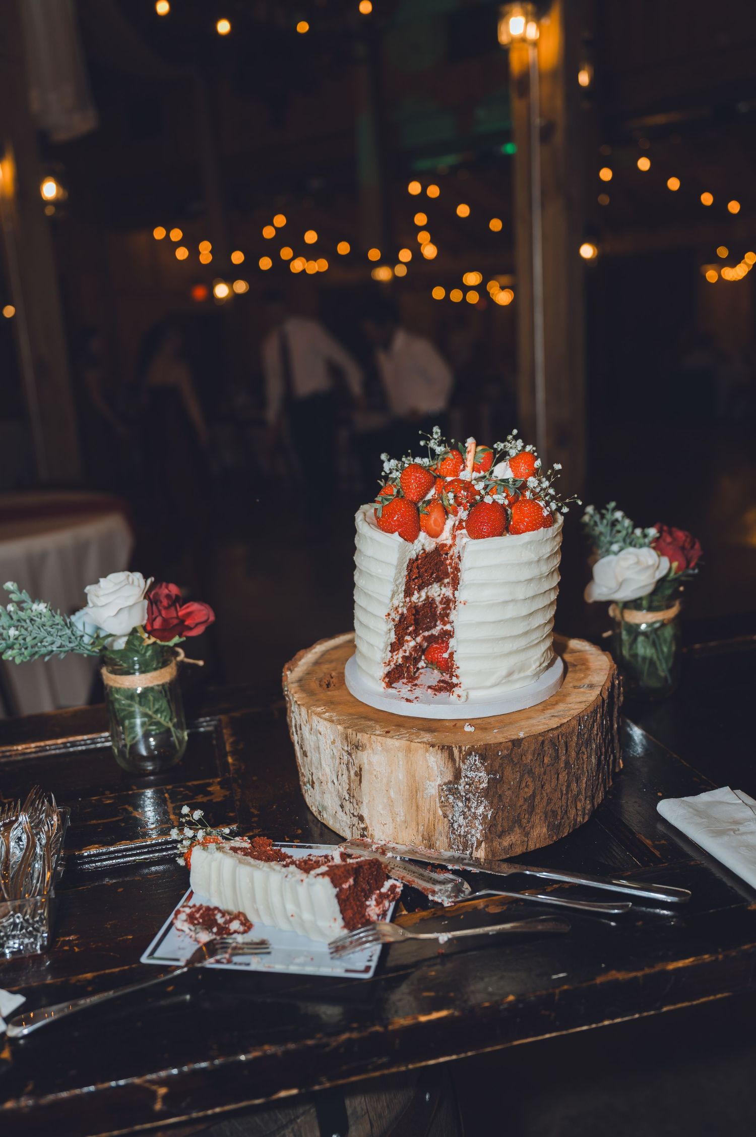 Single-tier red velvet wedding cake topped with strawberries on rustic wood stand, surrounded by floral arrangements and string lights.