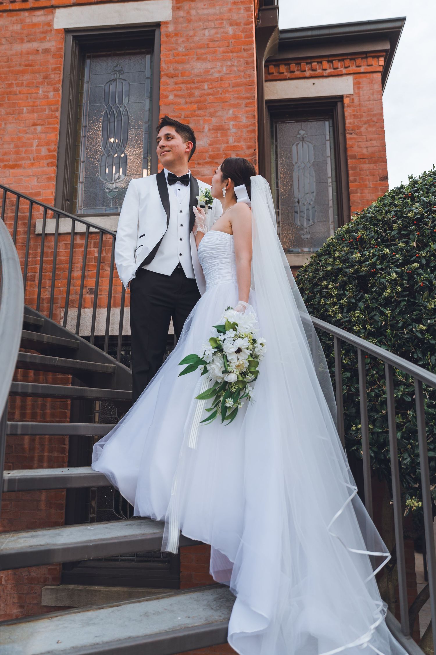 Bride and groom pose on brick building steps, she holds white bouquet with veil flowing, he wears white tuxedo jacket.