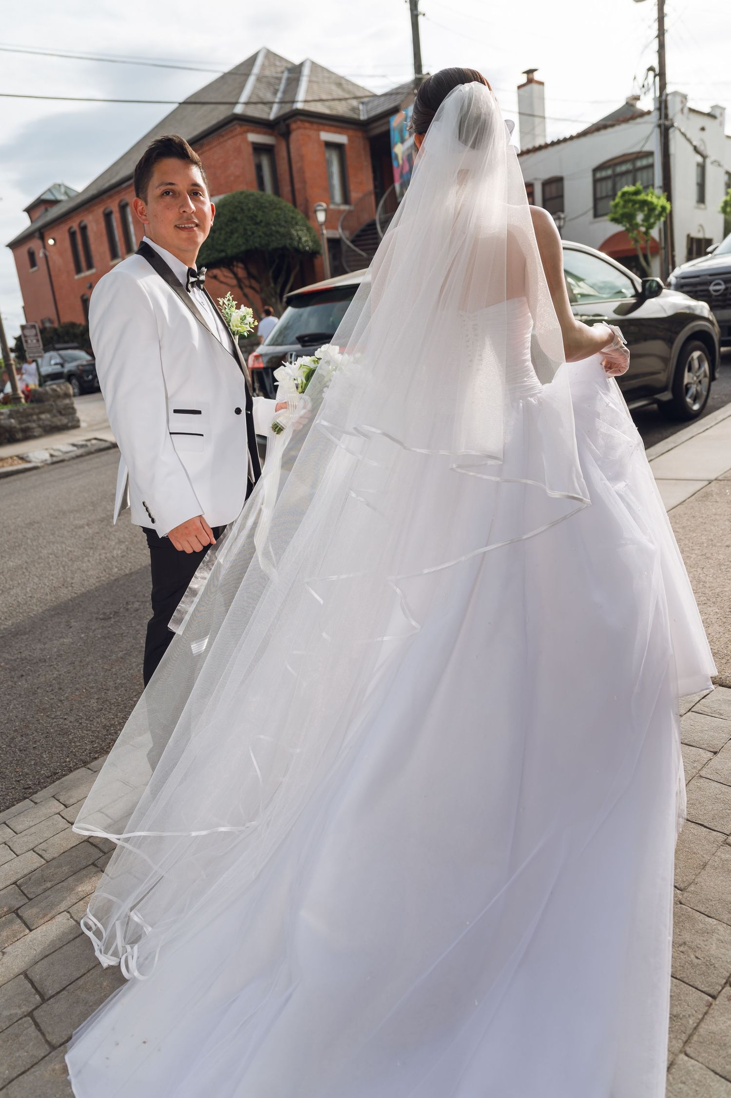 Bride in a flowing white gown with long veil walks with groom in white tuxedo on a sunny street.