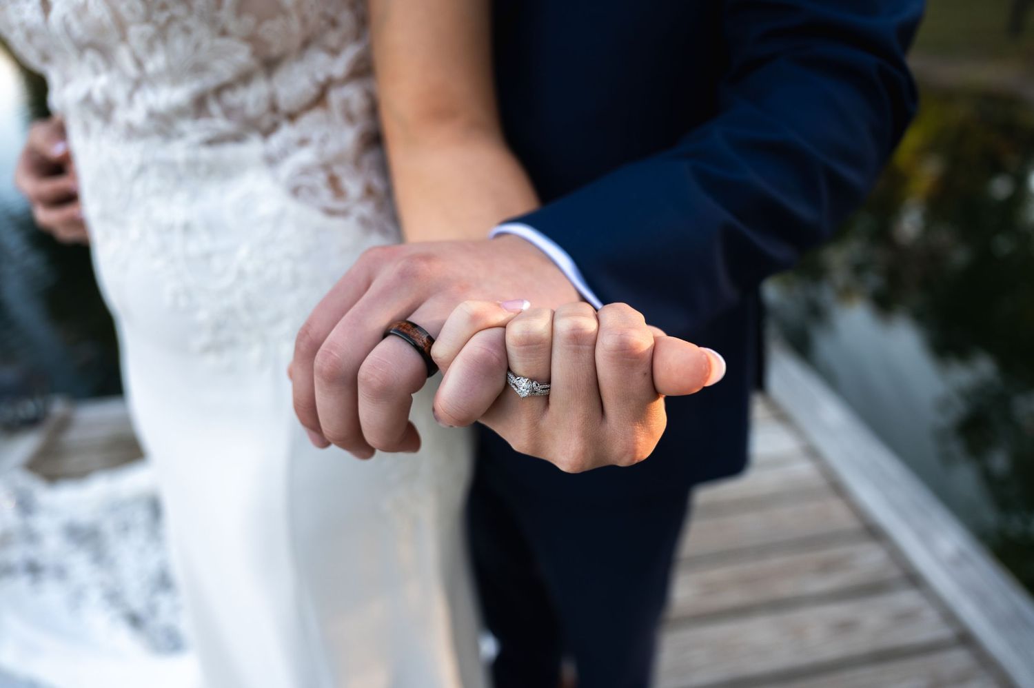 Newlywed couple holding hands showing wedding rings, bride in lace dress and groom in navy suit on wooden dock.