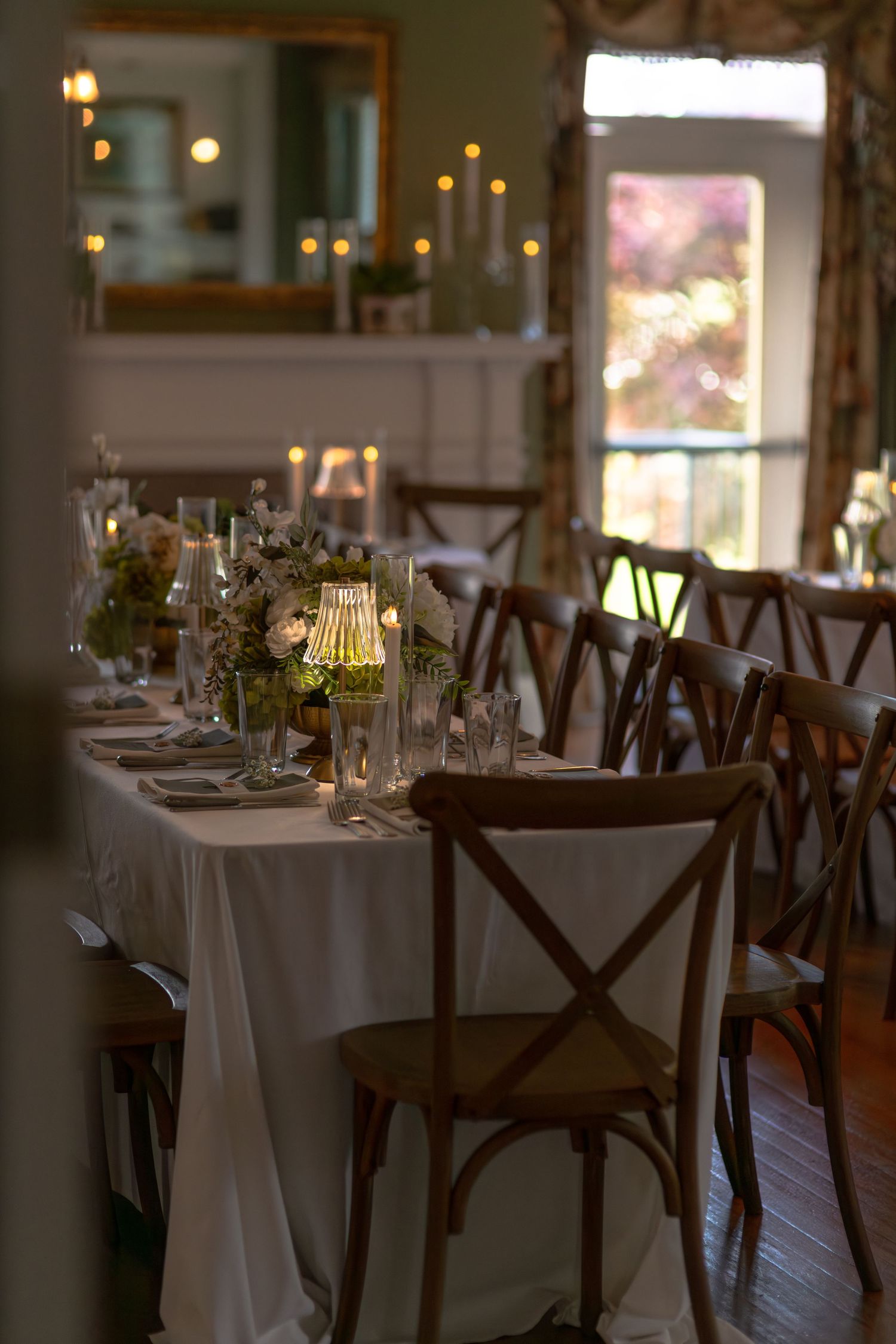 Elegant wedding reception table set with white linens, floral centerpieces, candles, and cross-back wooden chairs in a warmly lit room.