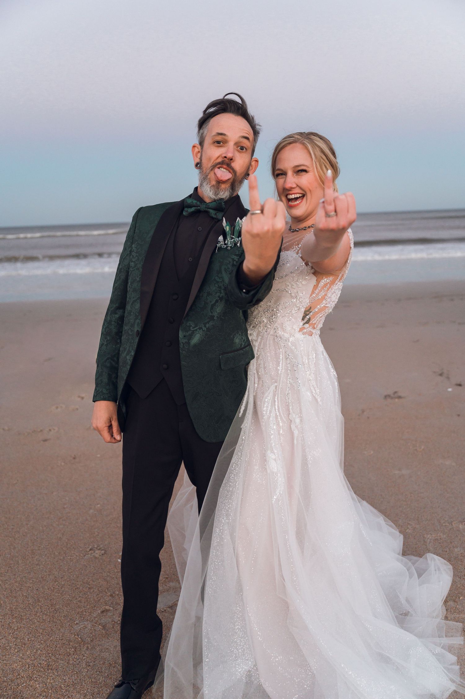 Newlyweds in wedding attire show off rings on beach, groom in green velvet blazer, bride in white lace gown.