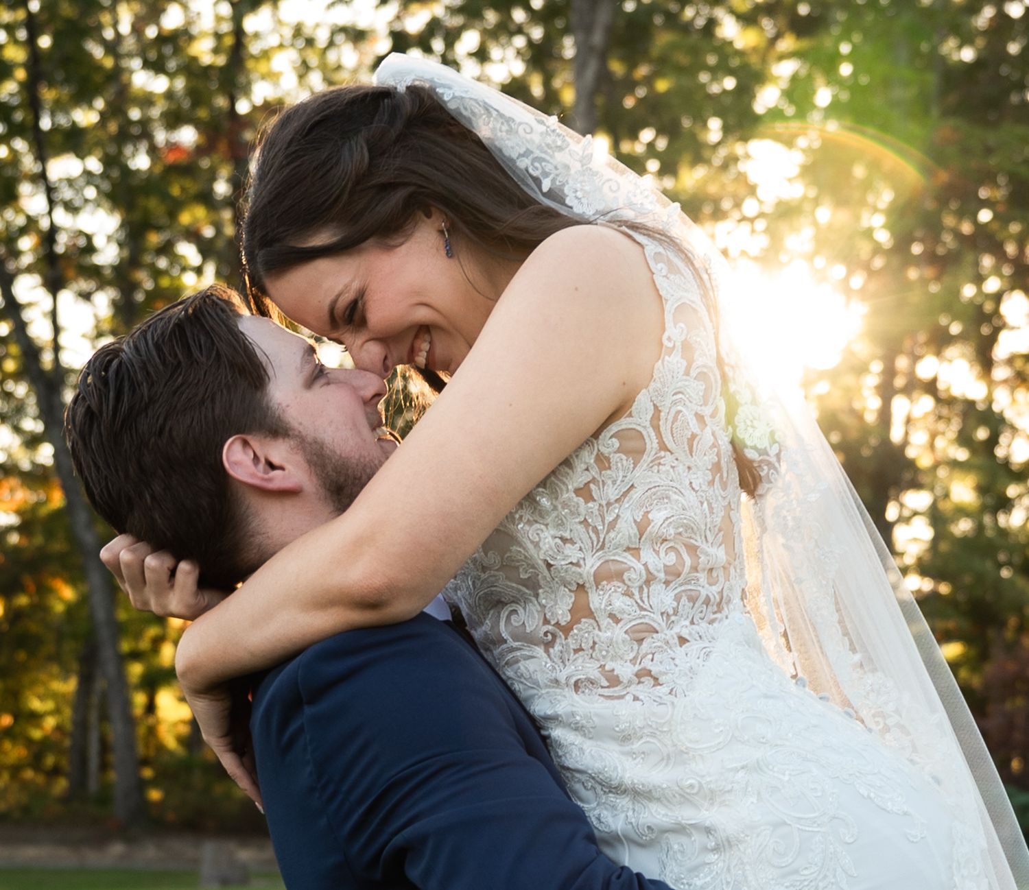 A groom lifts his smiling bride in a lace wedding dress outdoors, with golden sunlight filtering through trees.