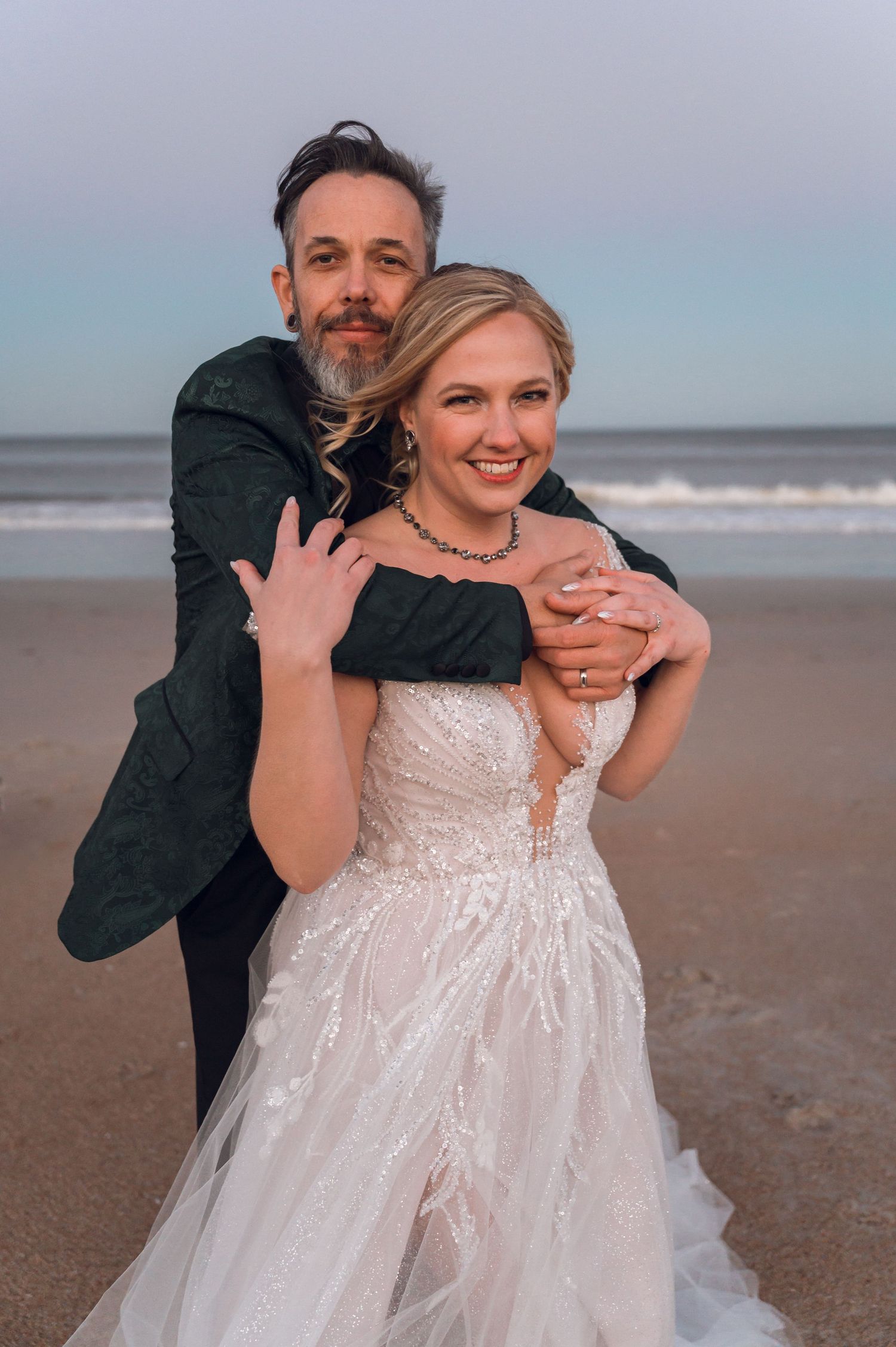 Bride in white lace gown embraced by groom in dark suit on a beach at dusk, smiling couple wedding photo.