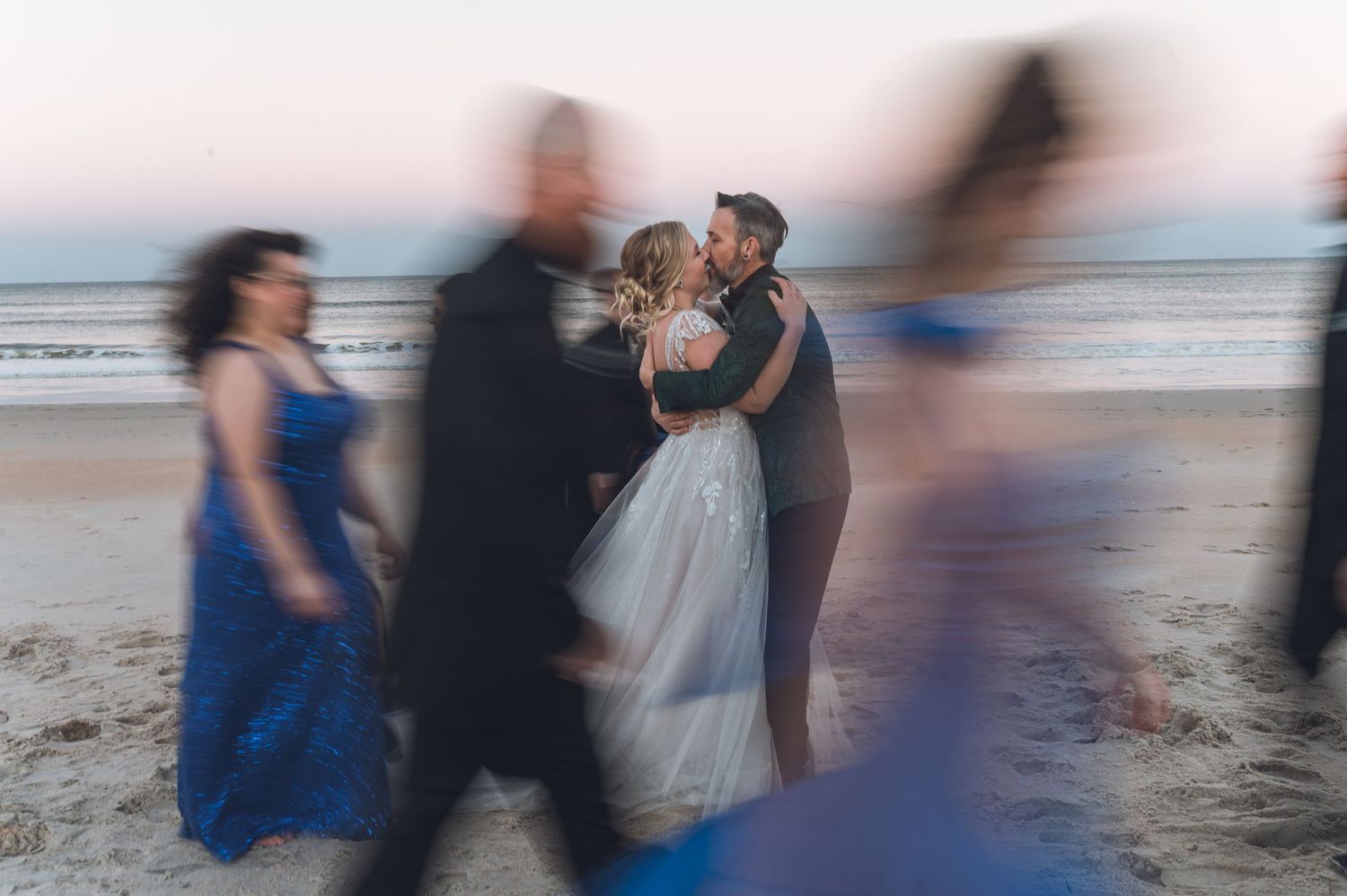 Couple shares a romantic beach wedding kiss at sunset, surrounded by bridesmaids in blue dresses.