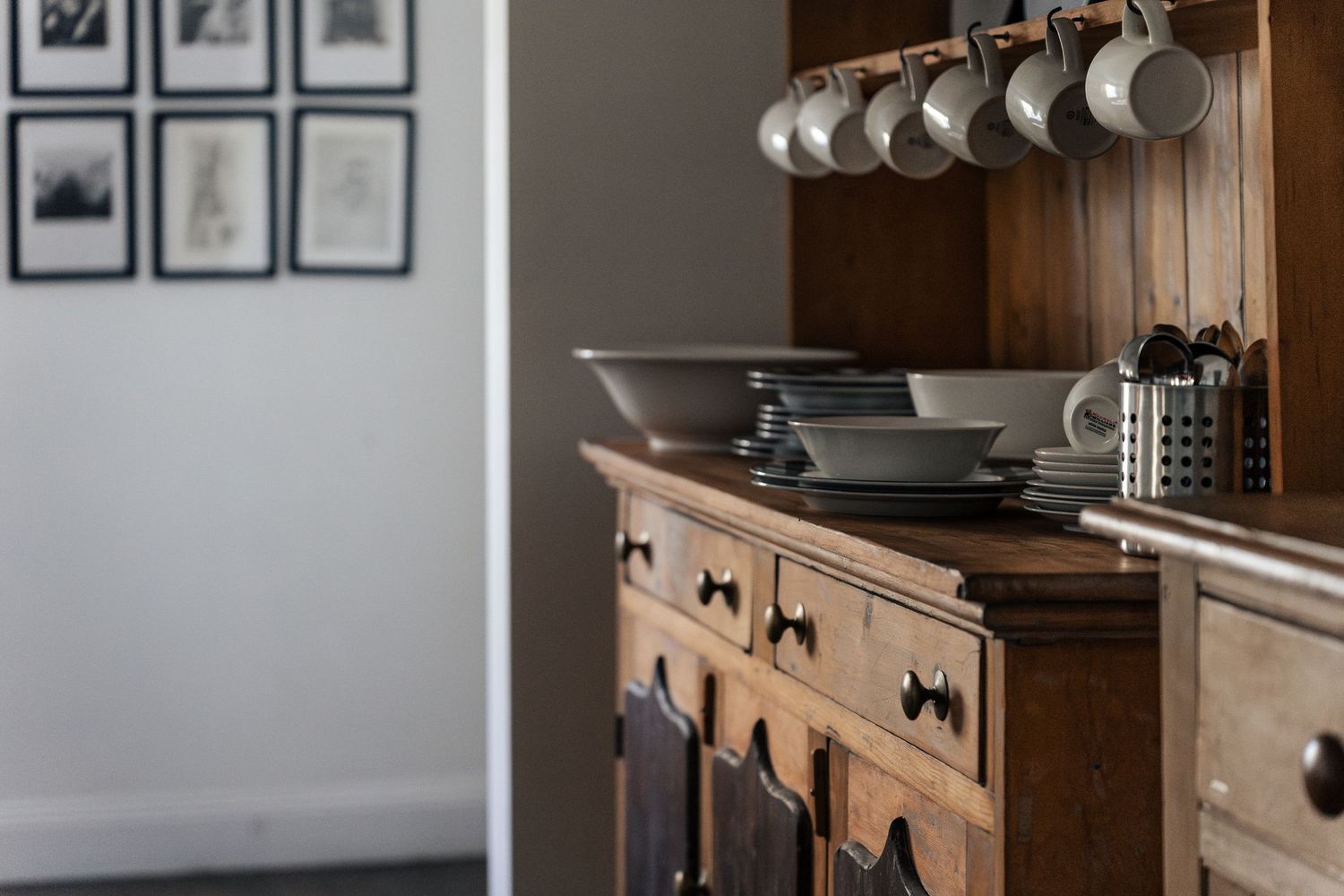 Rustic wooden sideboard with stacked grey dishes, hanging mugs, and framed black and white photos on a grey wall.

Piccadilly House