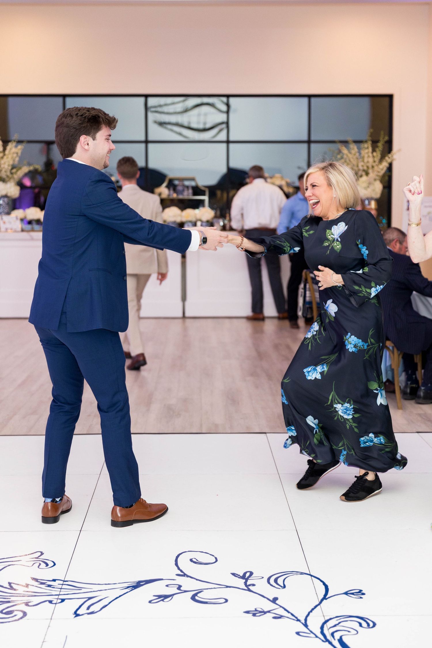 A young man in a navy suit dances joyfully with a woman in a floral dress at an elegant wedding reception.