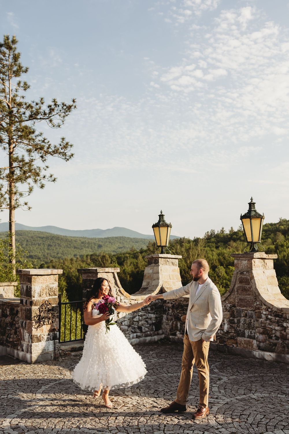 Bride and groom hold a veil on a stone terrace with mountain views and lanterns on a sunny wedding day.