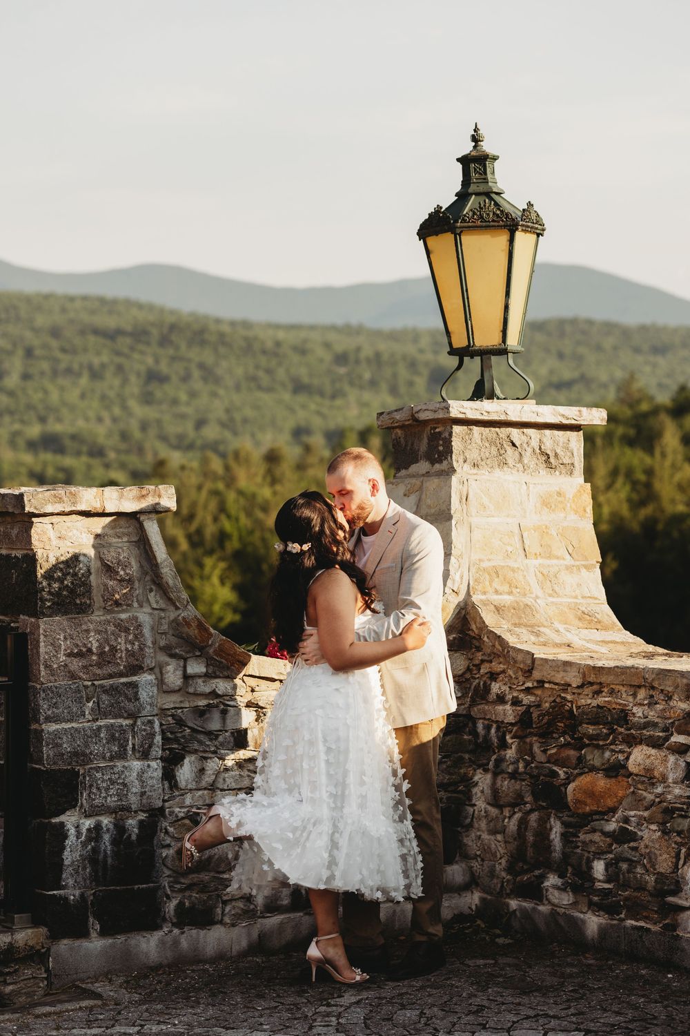 Couple sharing a romantic kiss on a stone terrace with a vintage lantern and lush green mountains in the background.