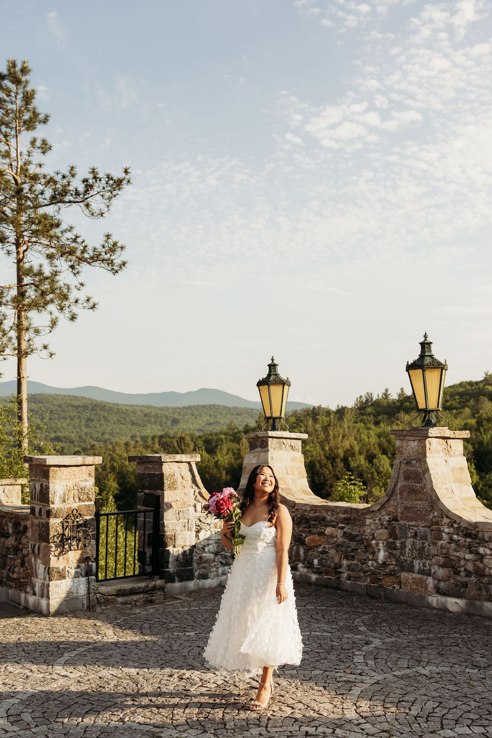 Smiling bride in white gown holds pink bouquet on stone terrace with mountain views and vintage lanterns.