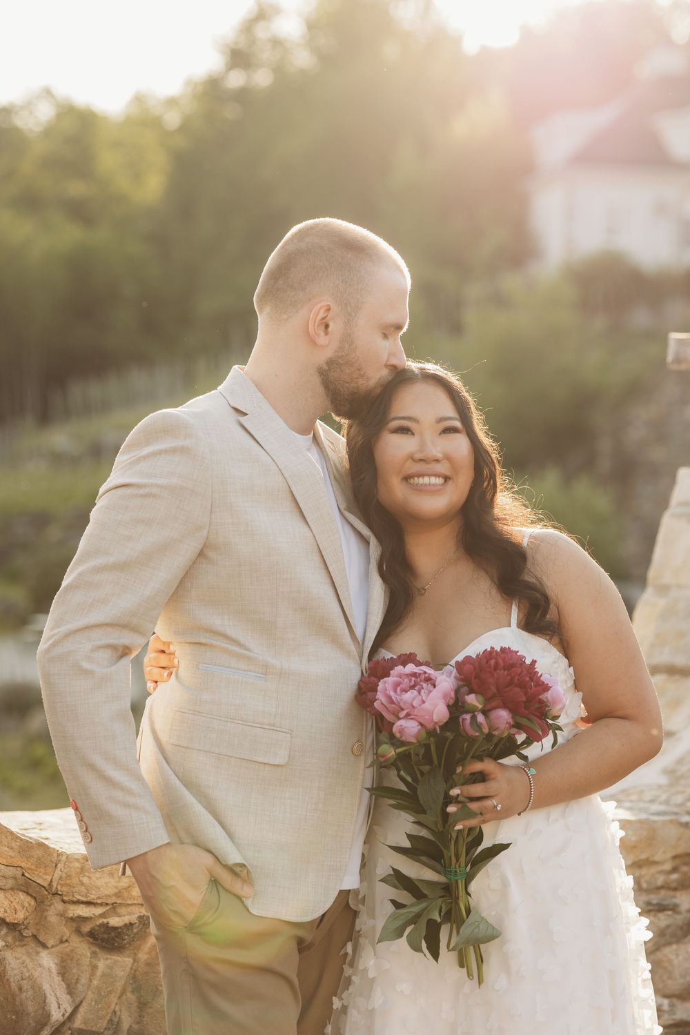 Bride and groom share a tender moment at golden hour, she holds pink peonies in a lace gown as he kisses her cheek.