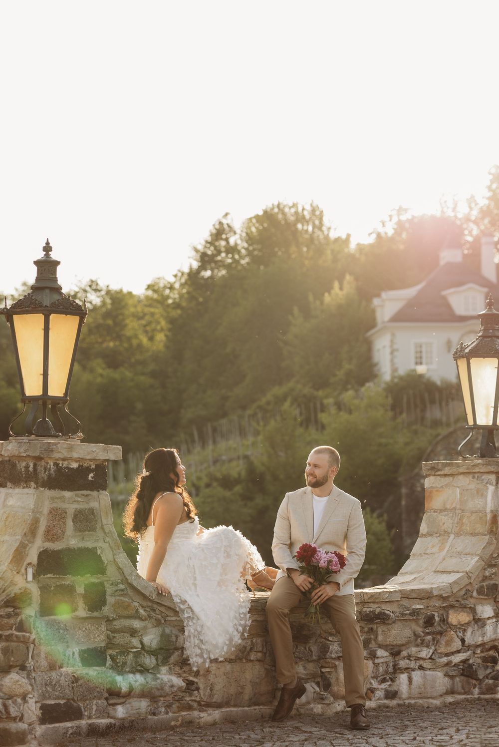 Bride and groom sit on stone wall flanked by glowing lanterns at golden hour sunset wedding.