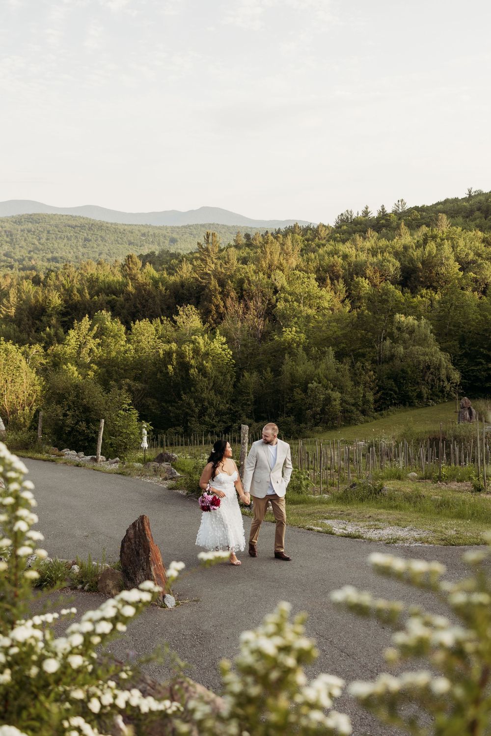Bride and groom walk hand in hand down a scenic path with lush green mountains in background.