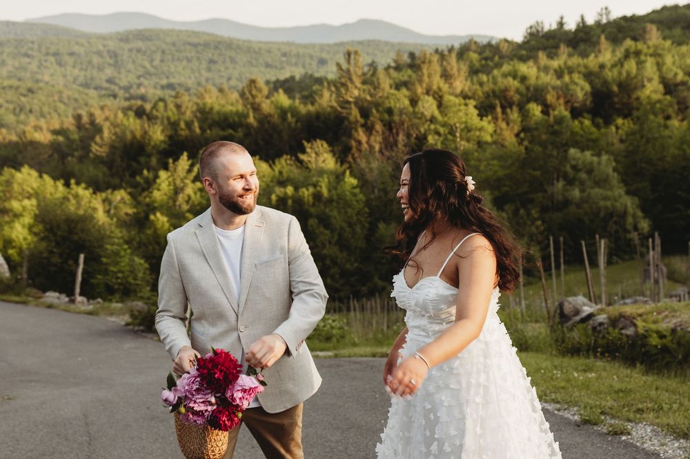 Bride and groom walking outdoors with mountain backdrop, holding pink bouquet, wearing wedding attire at golden hour.