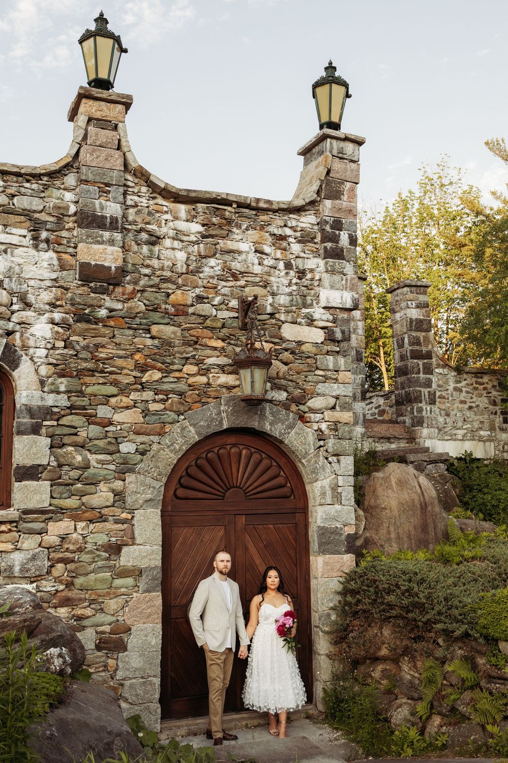 Bride and groom stand before rustic stone chapel arched wooden door with lanterns, bride holding pink bouquet.