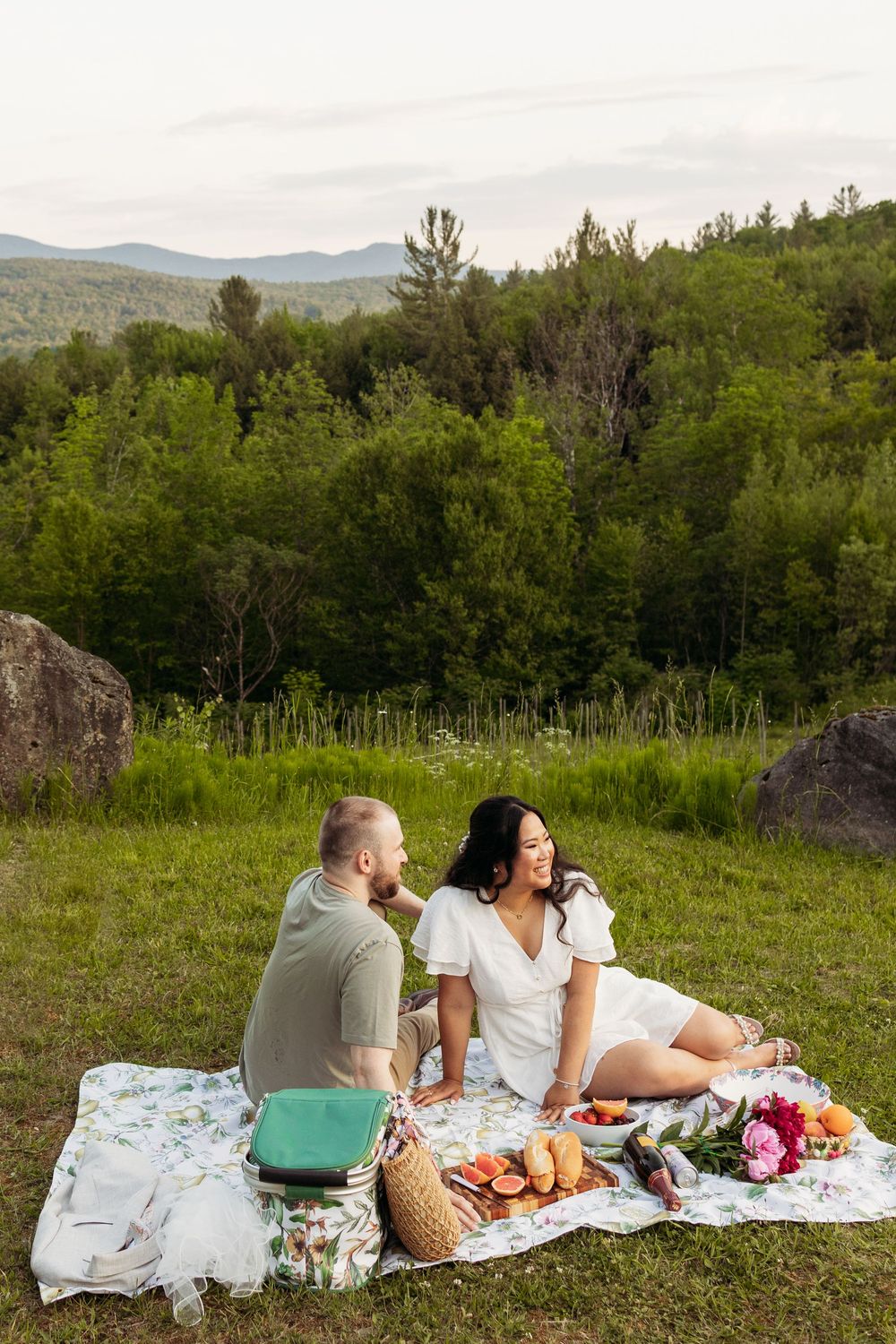 Couple enjoys a romantic outdoor picnic on a blanket with fruit and flowers, surrounded by lush green hills and trees.