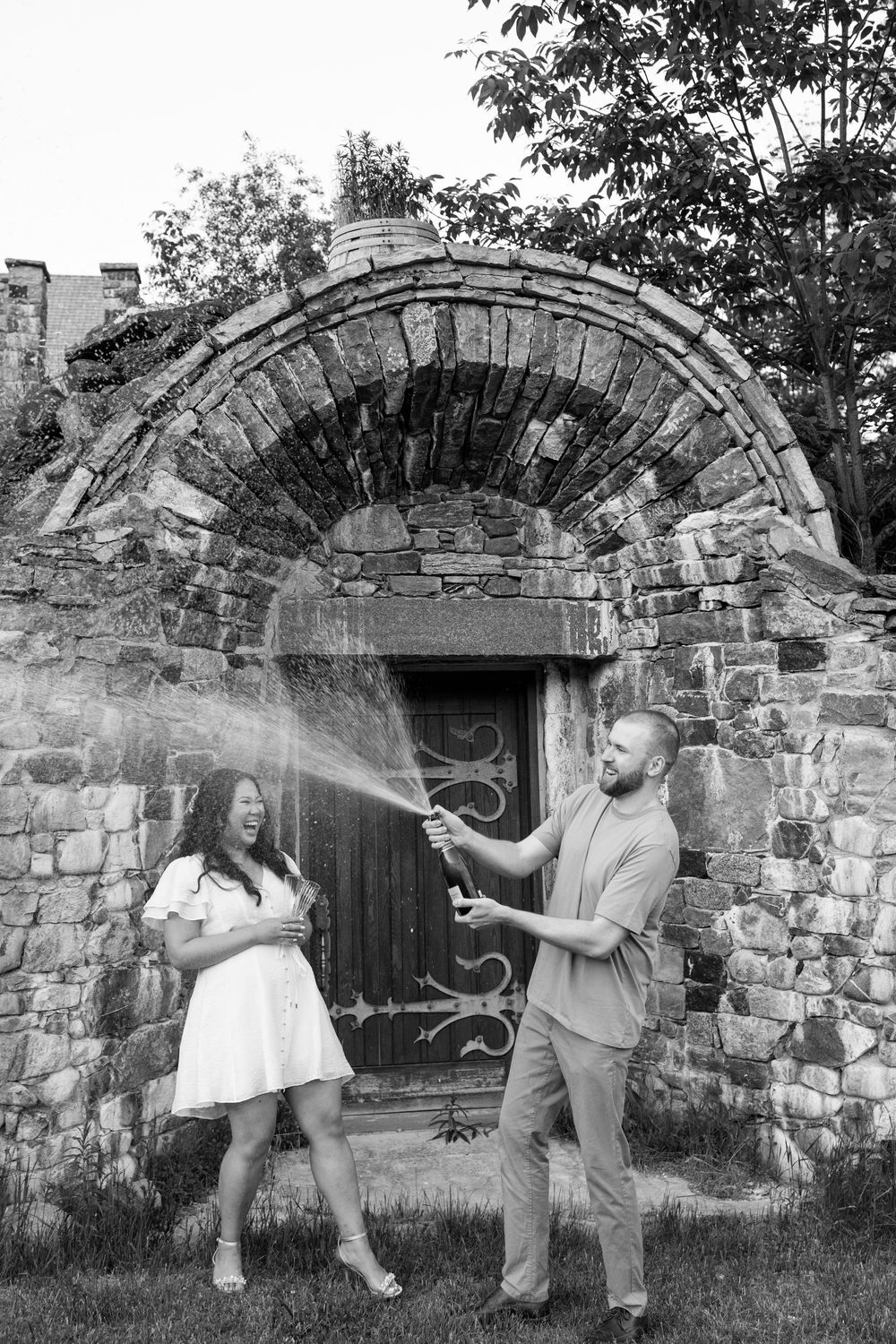 Groom pops champagne bottle spraying bride in white dress outside rustic stone arch doorway.