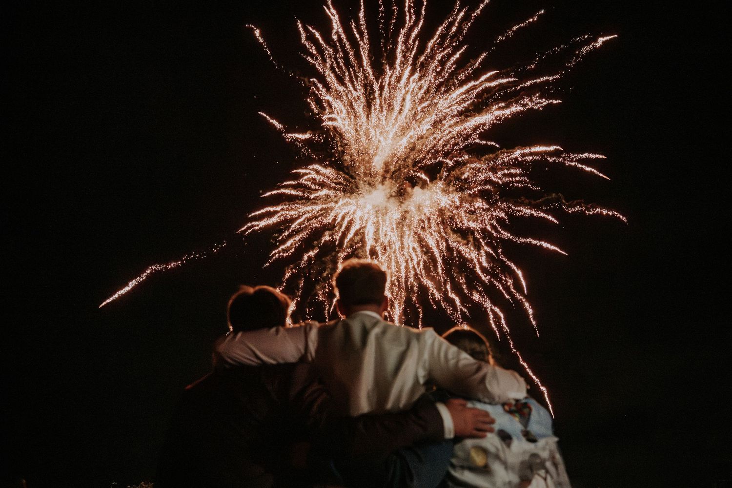 Three friends embrace while watching a stunning fireworks display burst in the night sky.