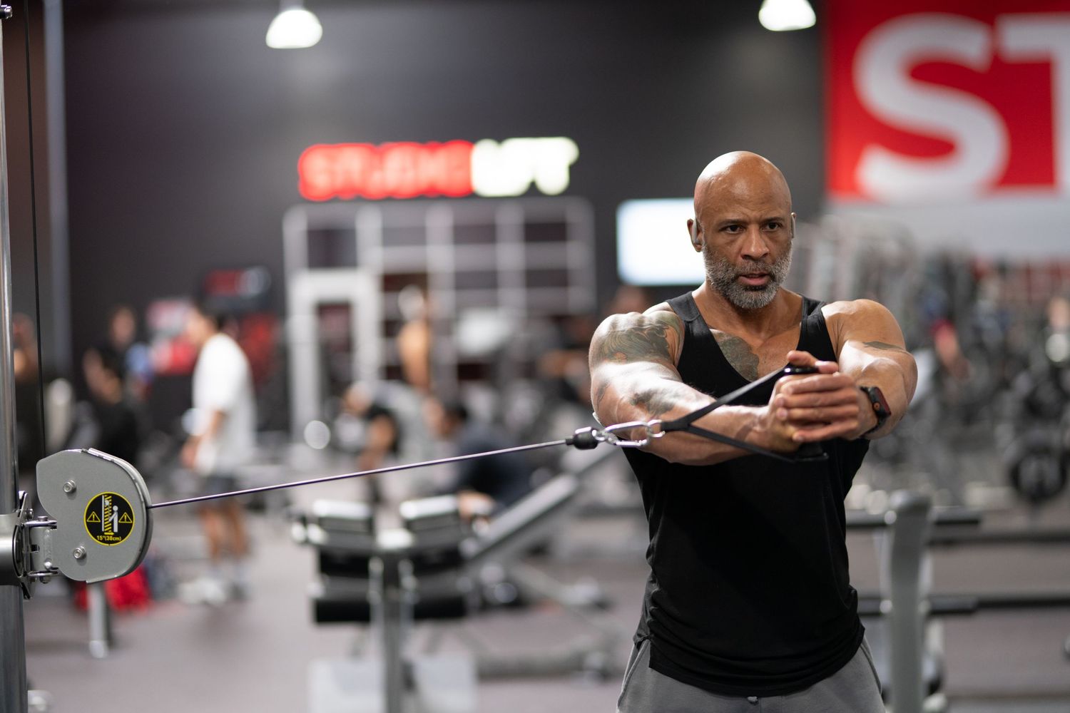 Muscular man performing cable crossover exercise at a modern gym, wearing a black tank top.
