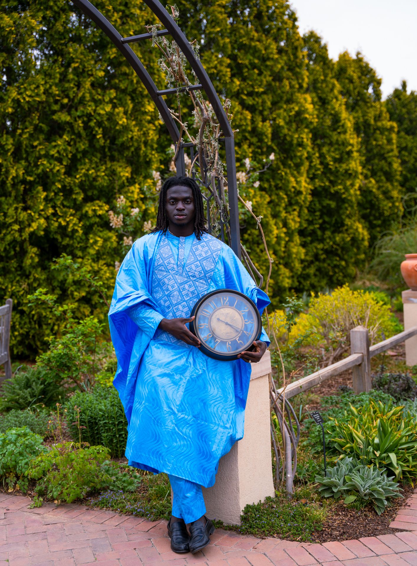 Man in blue traditional African attire holds a round clock while standing in a lush garden under a metal arch.