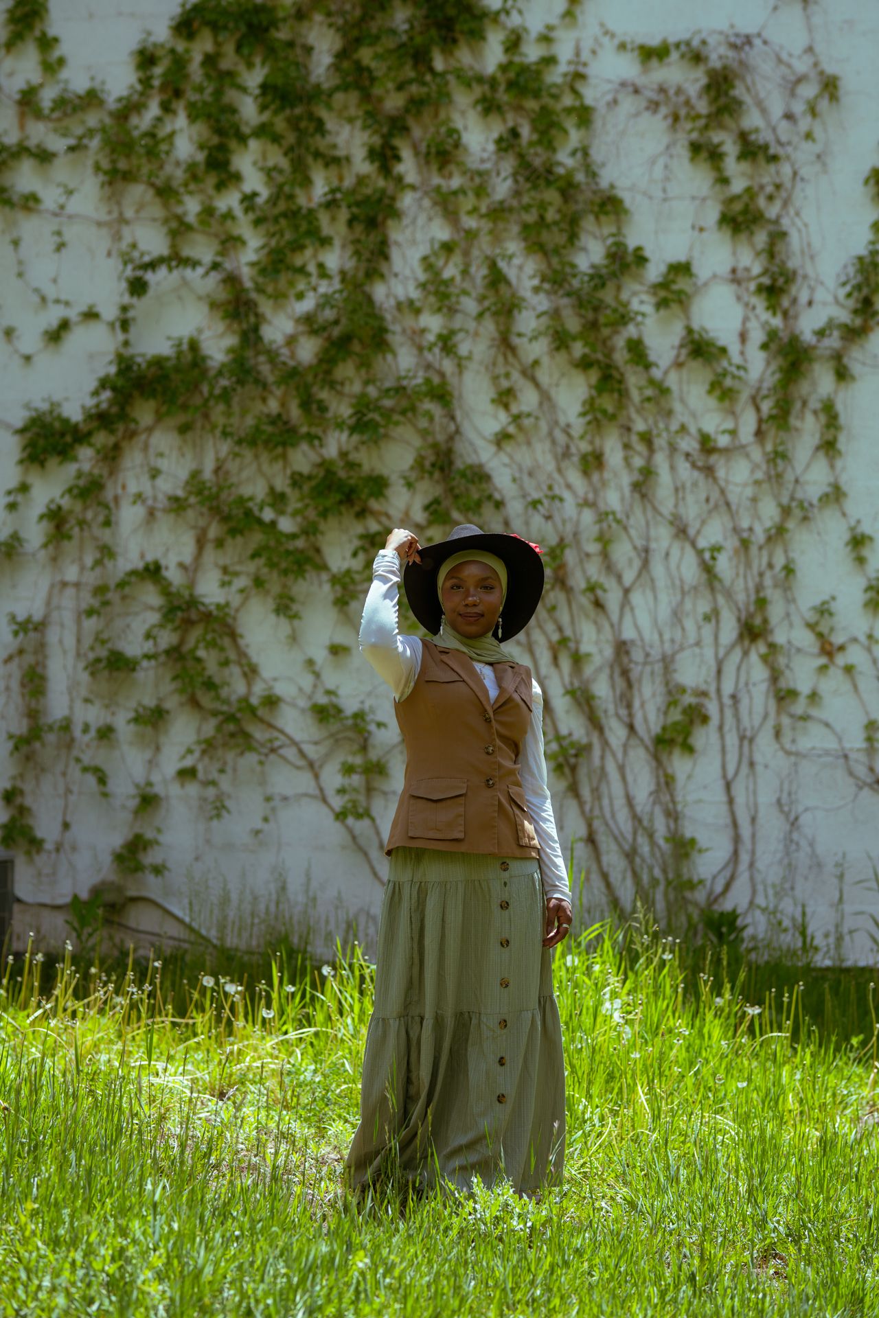 Woman in vintage outfit with wide-brim hat stands on green grass before ivy-covered white wall.