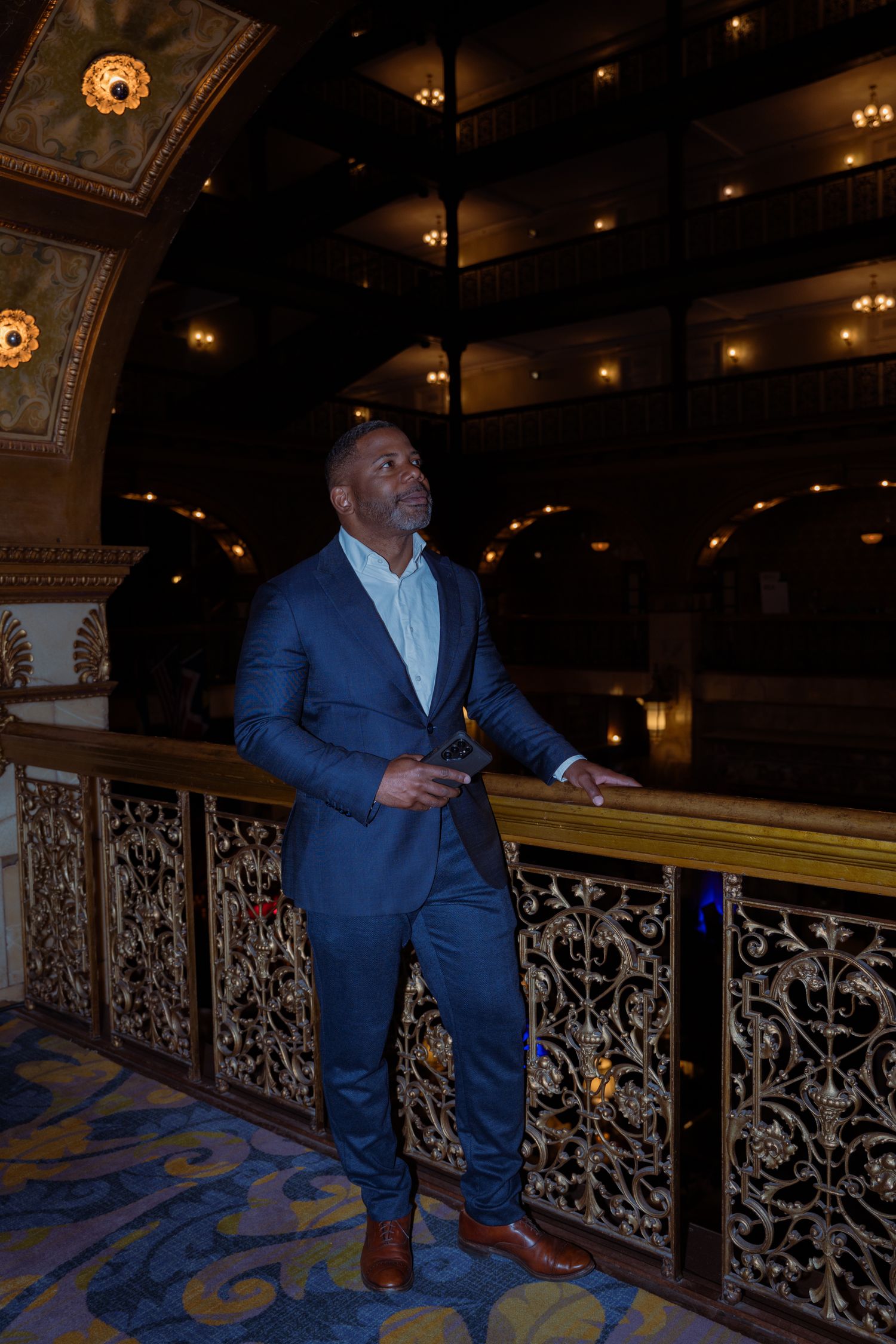 Elegant man in blue suit standing on ornate balcony inside grand theater with warm golden lighting.