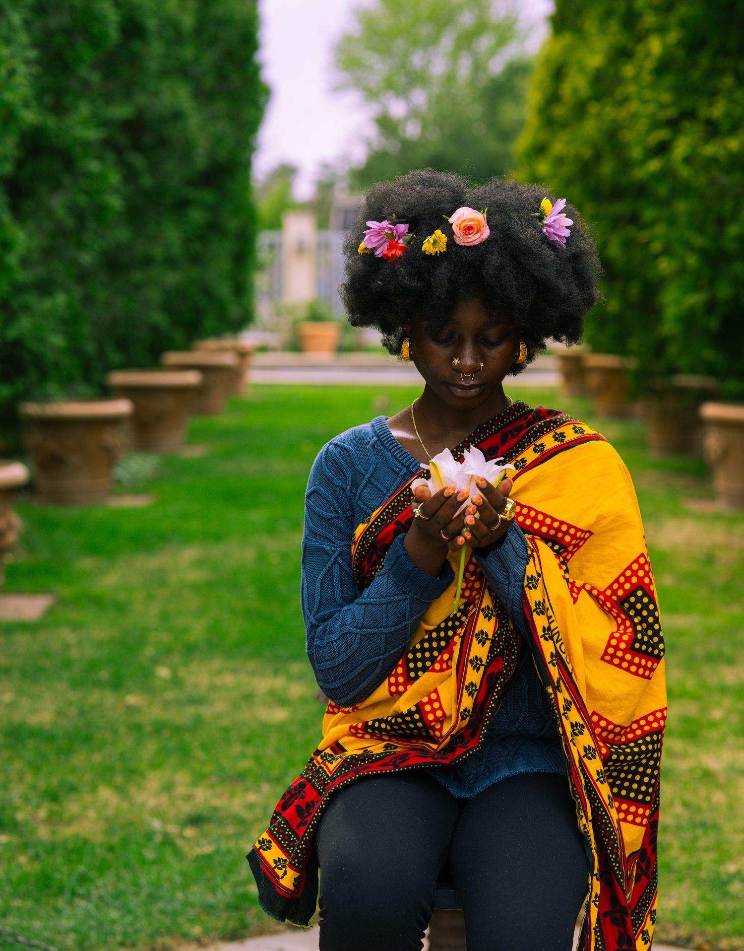 Young Black woman with flowers in afro hair, wearing colorful African print wrap, holding flower in lush garden.