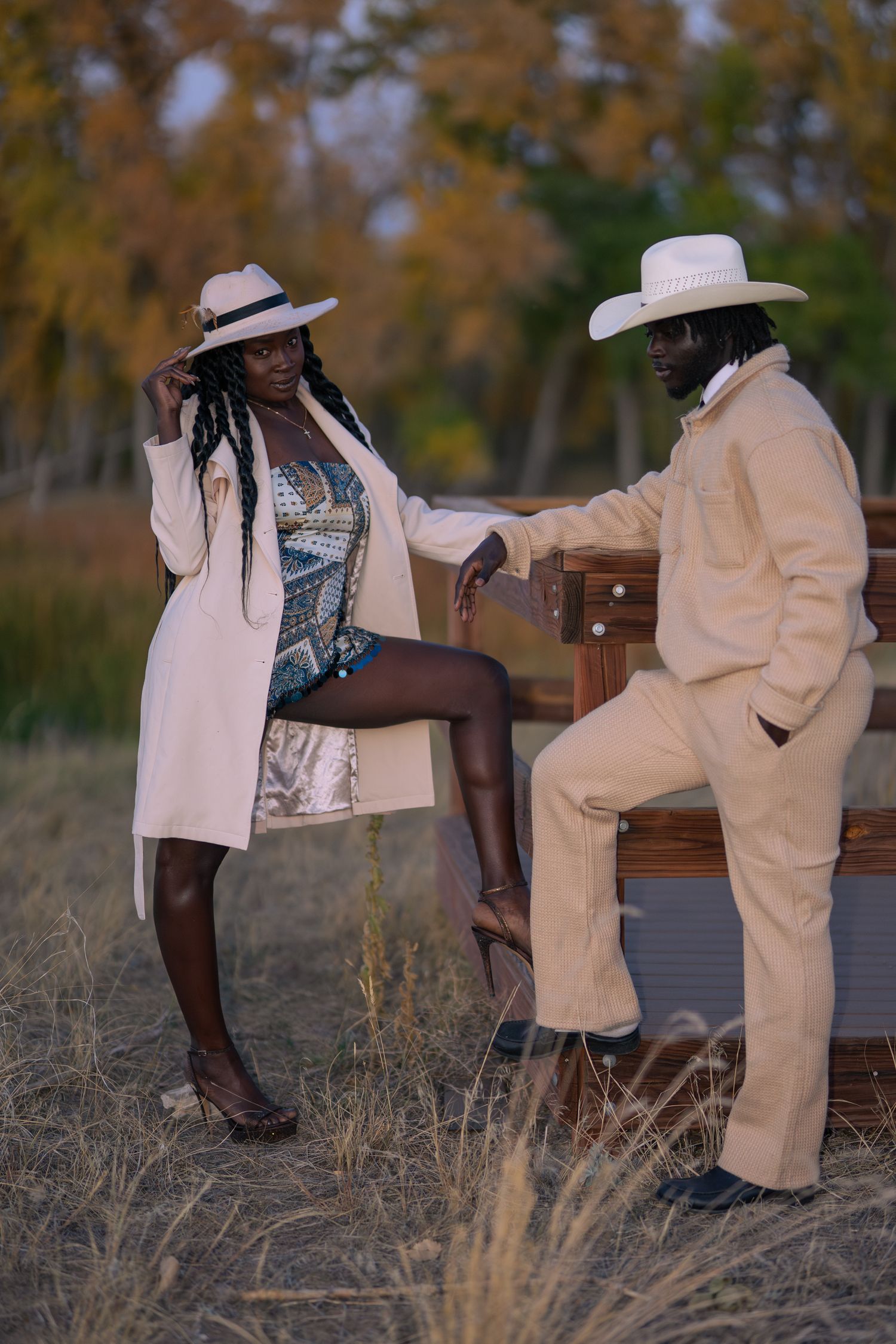 A stylish Black couple in matching cream outfits and cowboy hats pose outdoors in a field with autumn trees.