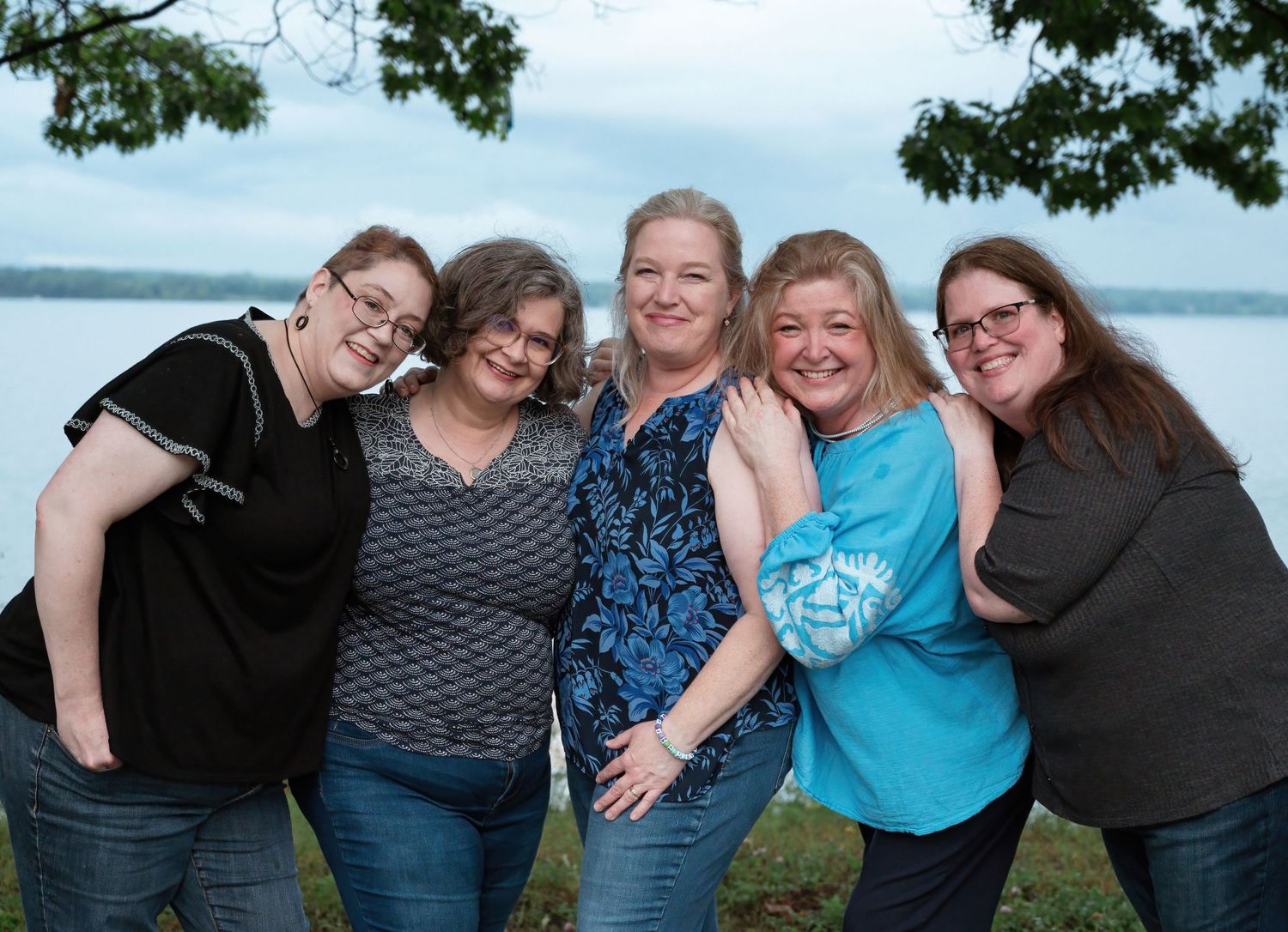 Five smiling women pose together outdoors near a lake on a cloudy day.