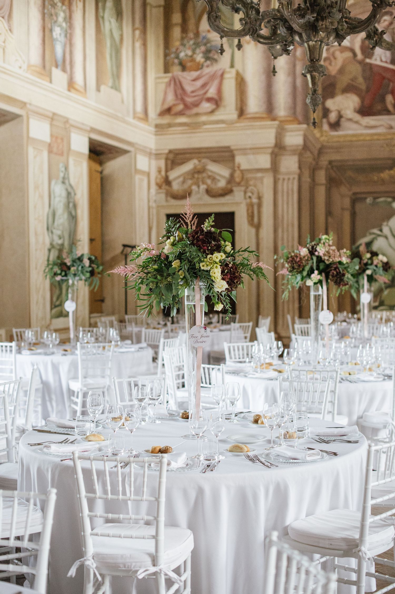 Elegant wedding reception in ornate villa with white round tables, chiavari chairs, and lush floral centerpieces.