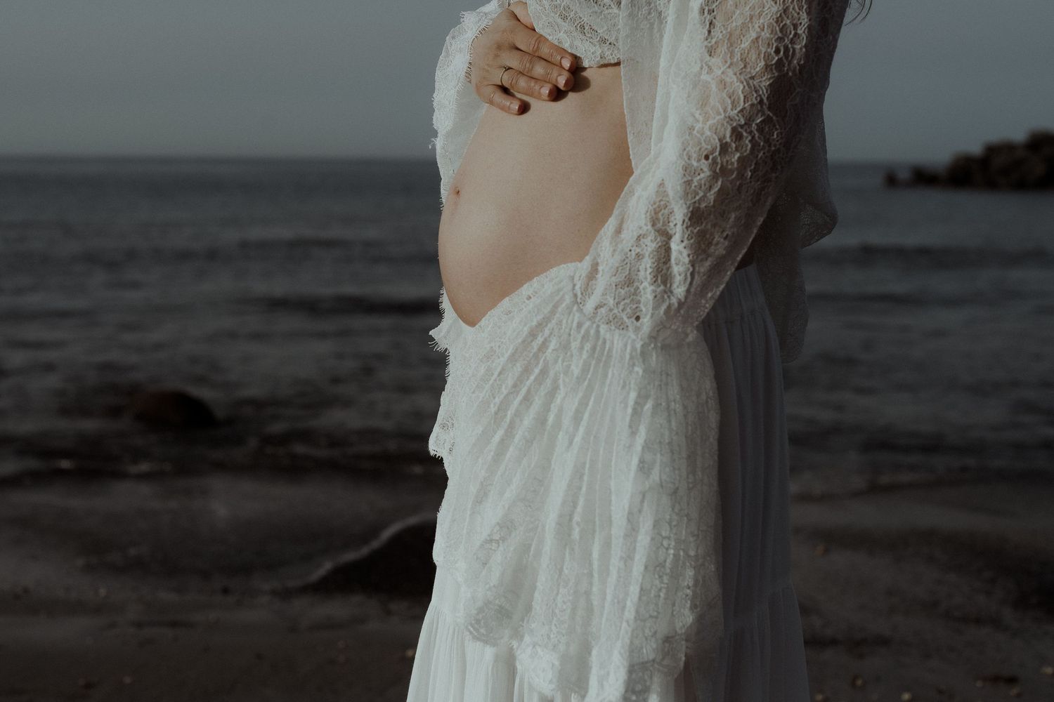 Pregnant woman in white lace dress standing by the ocean shore, cradling her baby bump at dusk.