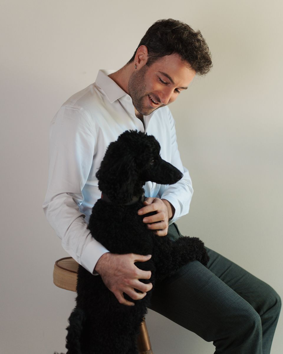 A smiling man in a shirt sits on a stool with a black poodle dog against a white background dog photoshoot in Sydney Hills district