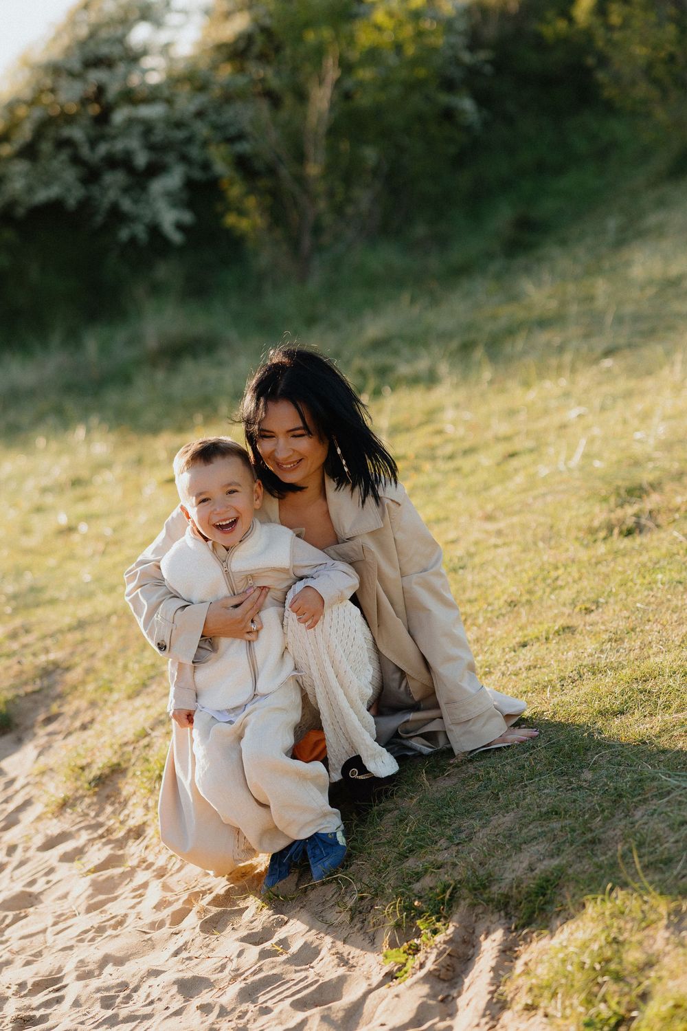 A smiling mother crouches on a sandy hill hugging her laughing toddler in white outfit outdoors.