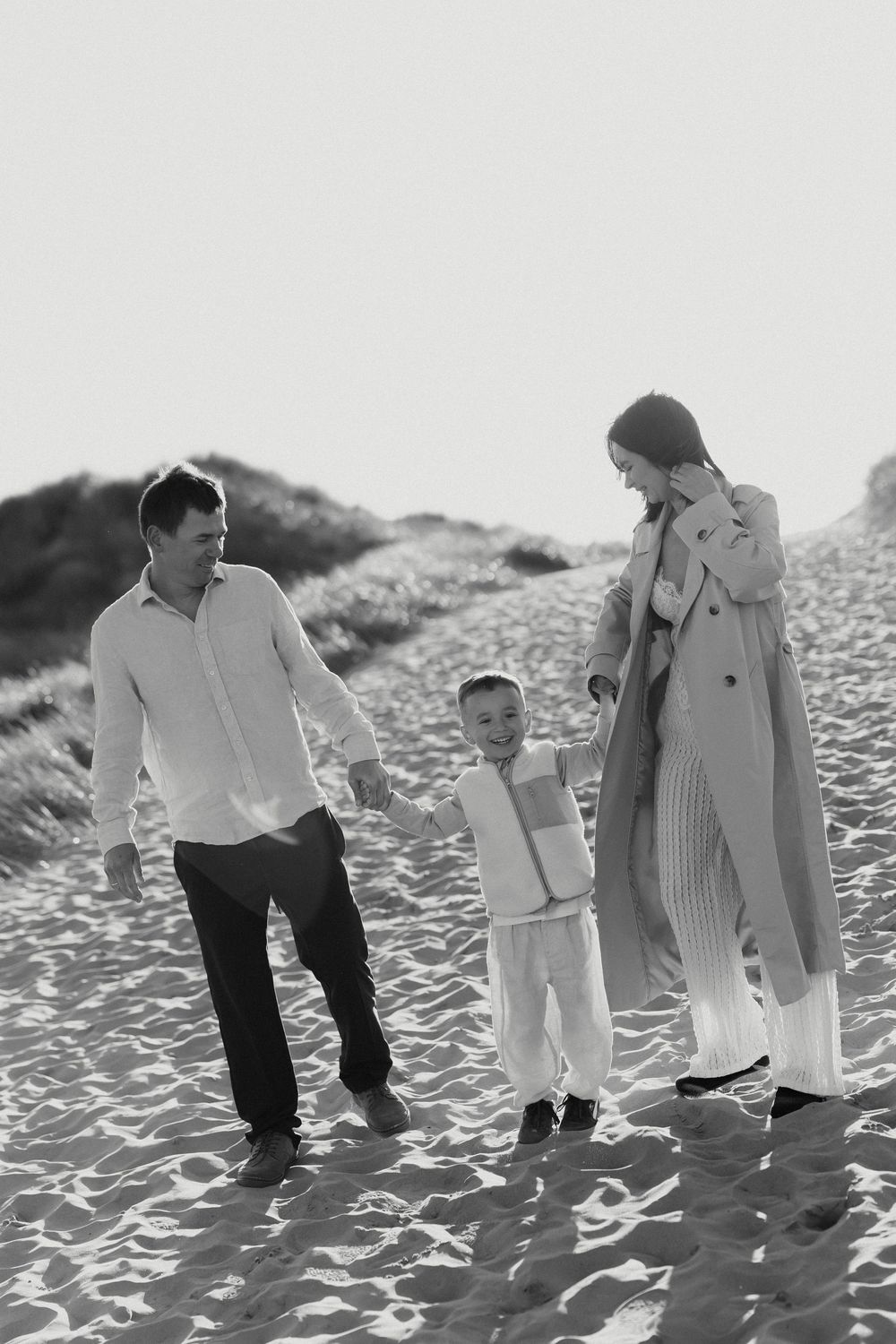 Black and white photo of a happy family walking on sandy beach dunes, parents holding child's hands.