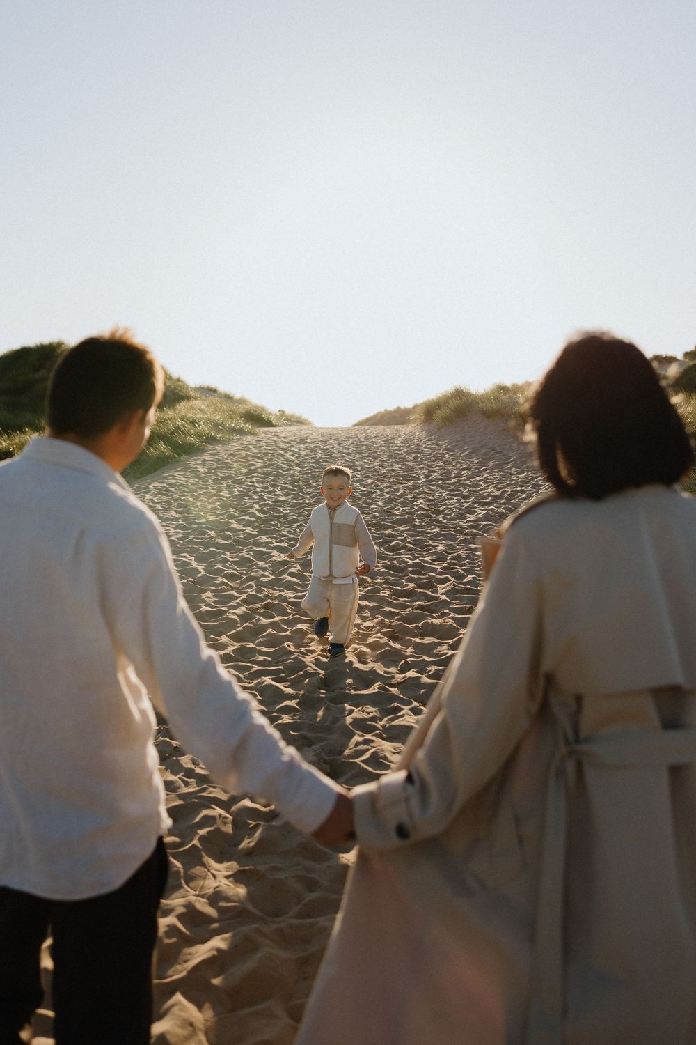 A child runs toward two adults holding hands on a sandy beach path, with grass dunes and bright sky in the background.
