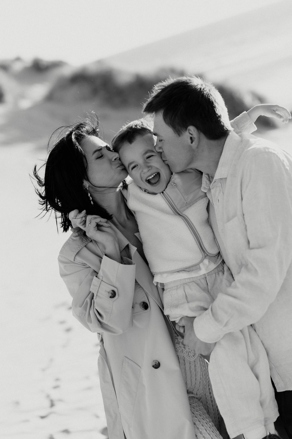 Black and white photo of parents kissing their laughing toddler on the cheeks outdoors on a sandy beach.