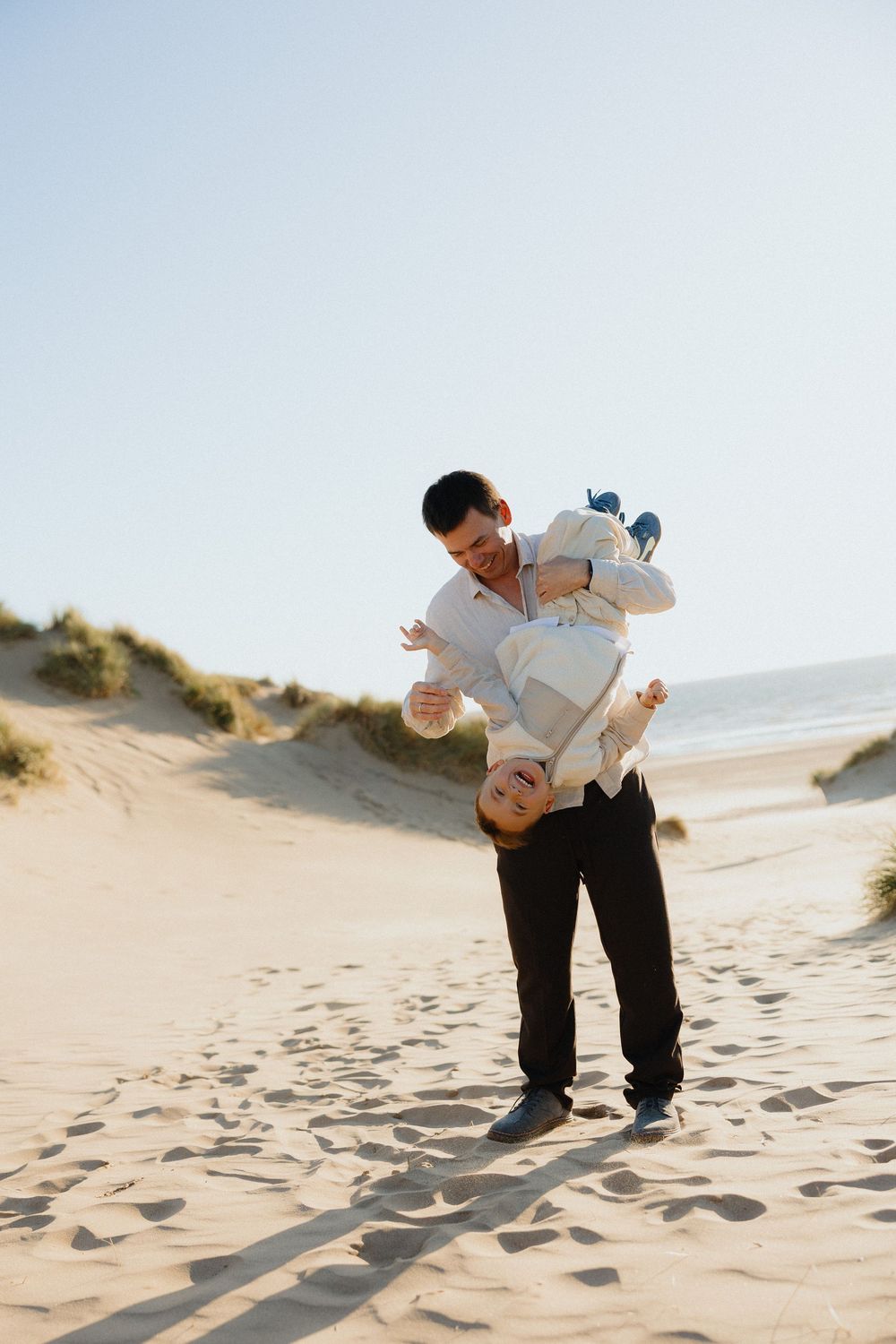 Father playfully holding young son upside down on sunny sandy beach.