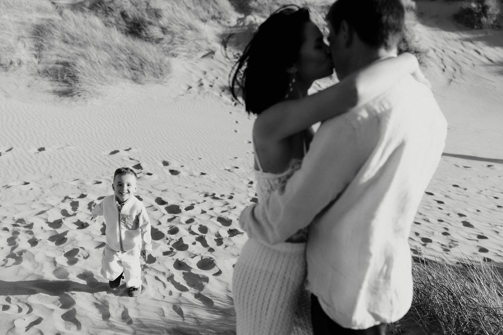 Black and white beach photo of a couple kissing while a young child stands in the sand behind them.