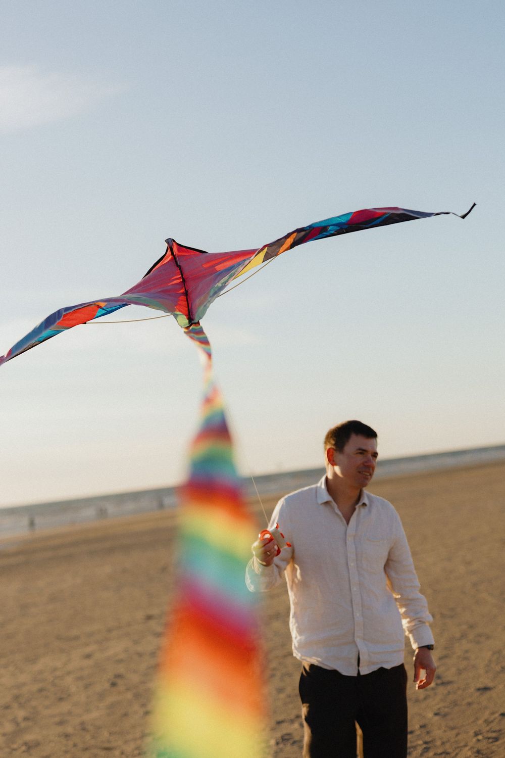 Man in white shirt flying a colorful rainbow kite on a sandy beach at sunset.