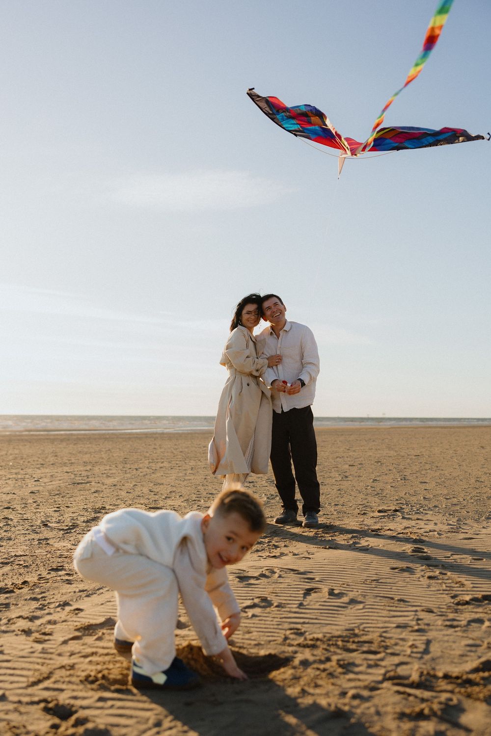 Family flying colorful kite on beach while young child plays in sand.