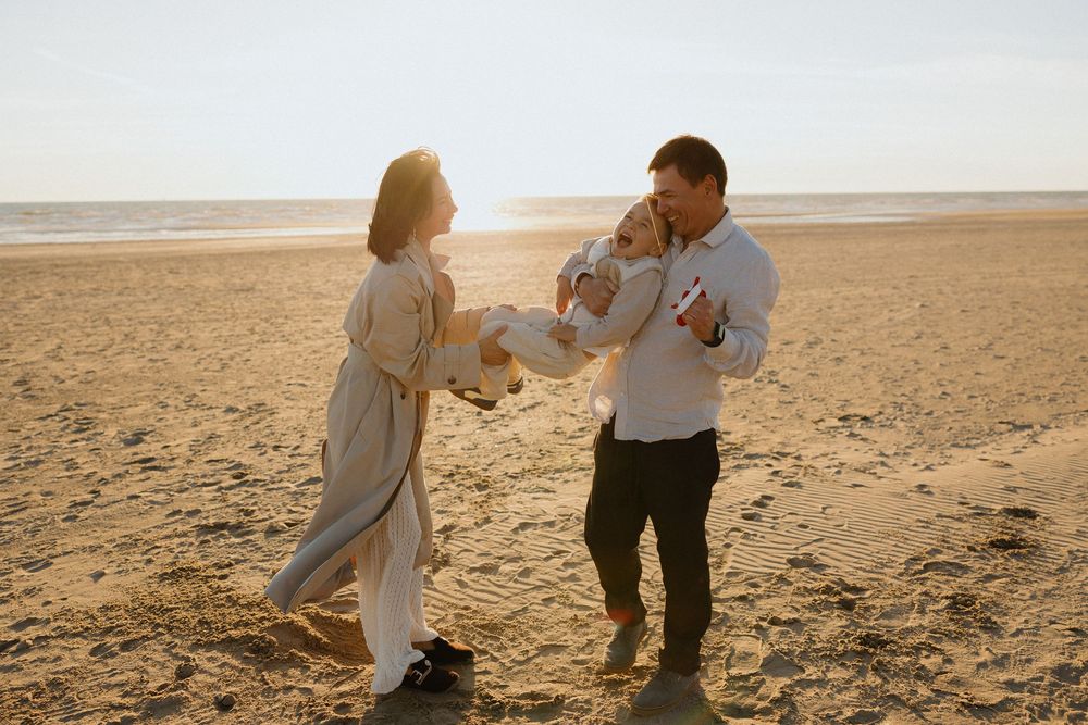 Family of three enjoying a sunny beach day, parents holding their toddler while smiling and playing on golden sand.