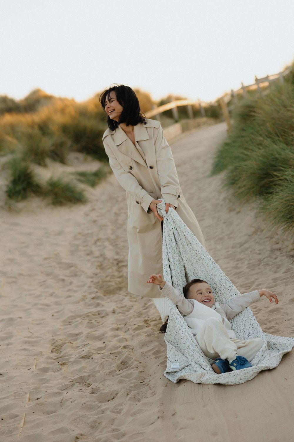 Mother dragging laughing child on blanket across sandy beach at sunset.
