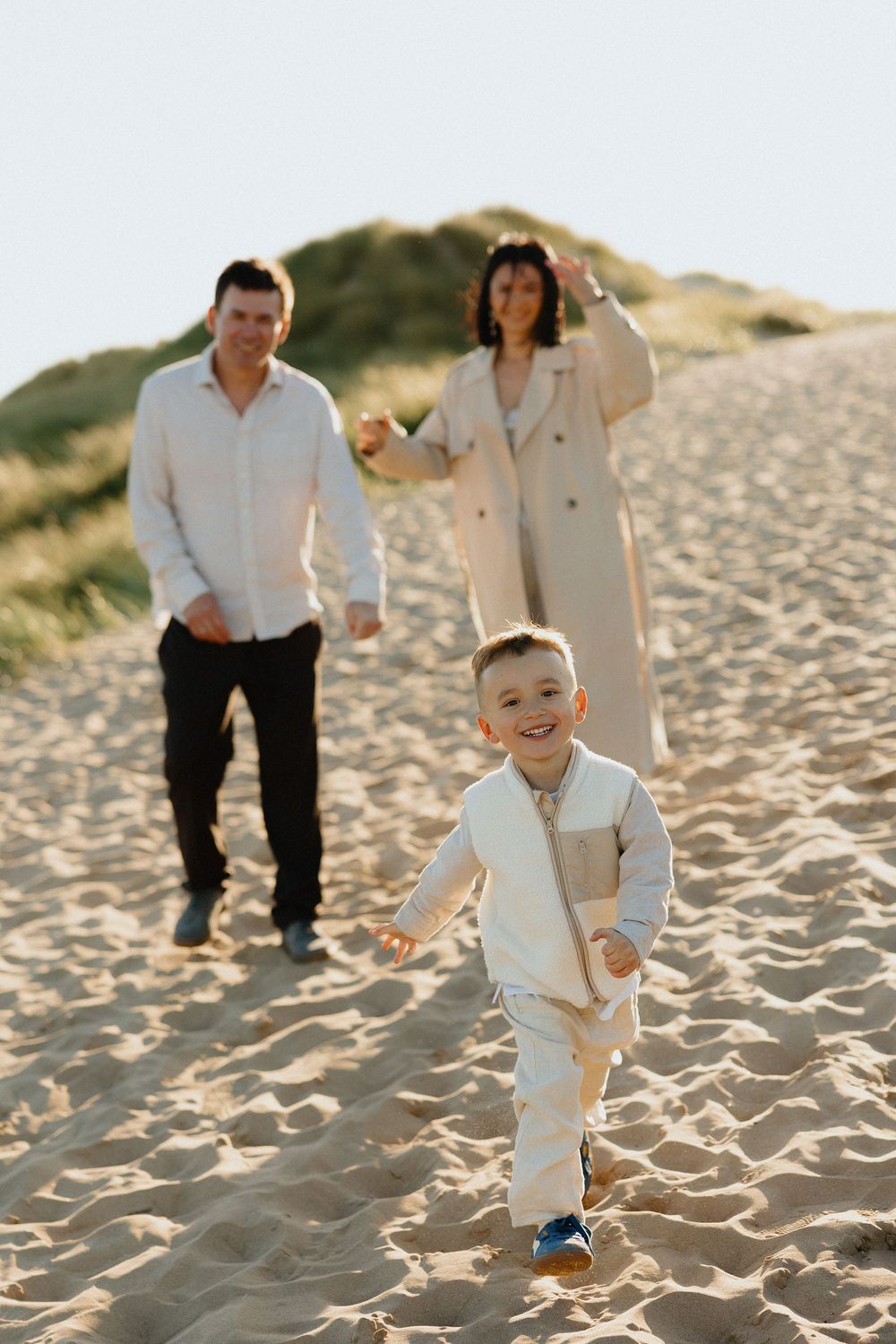 Happy family with young boy running on sandy beach dunes during golden hour.