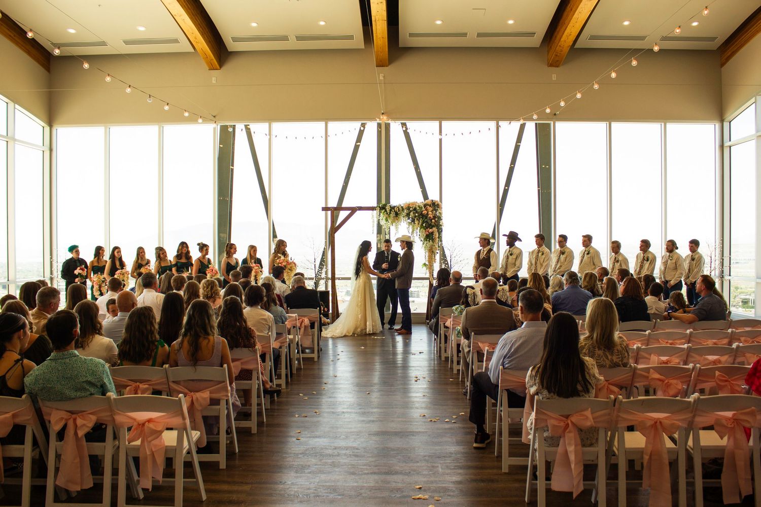 A bride in a white gown smiles at her groom in a cowboy hat near a sunlit window with floral decor.