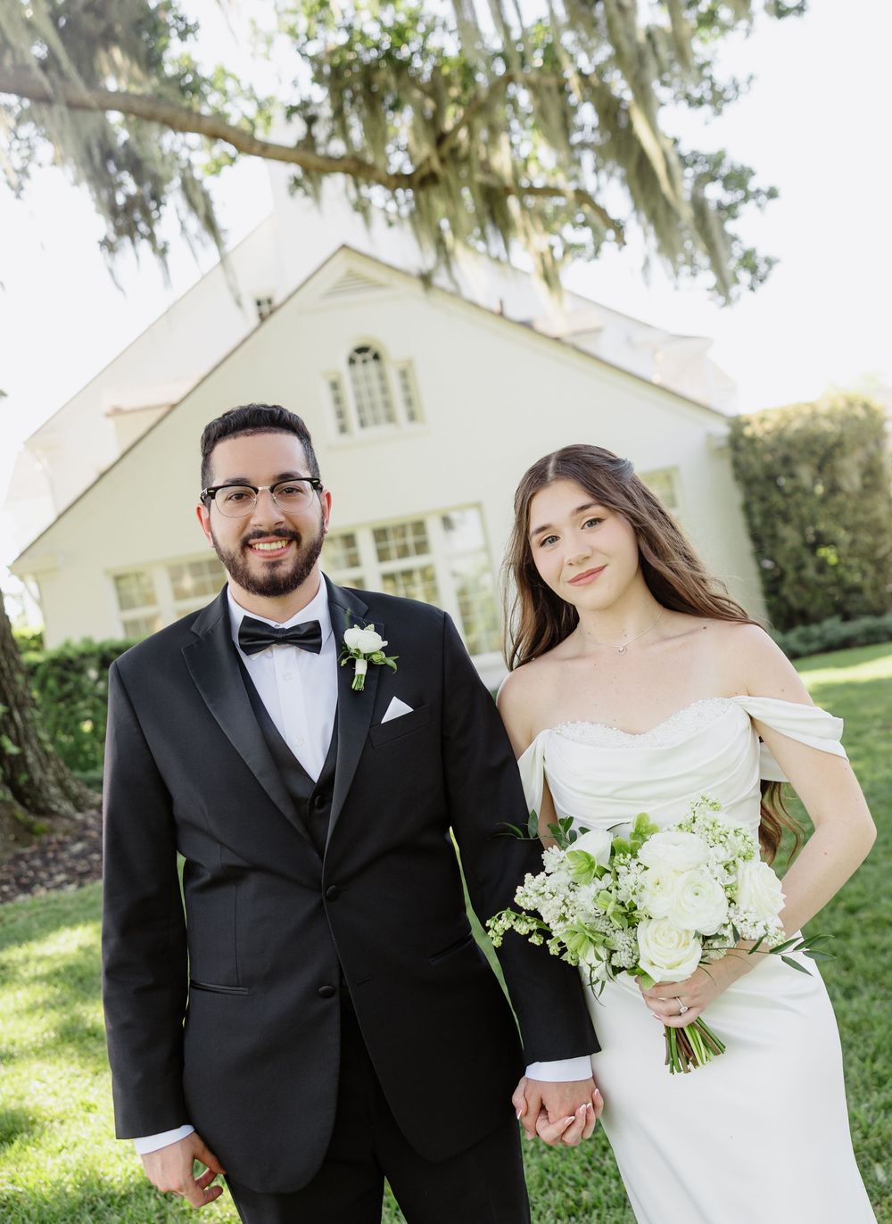 Bride and groom holding hands in front of a white chapel, bride holding white bouquet, groom in black tuxedo.