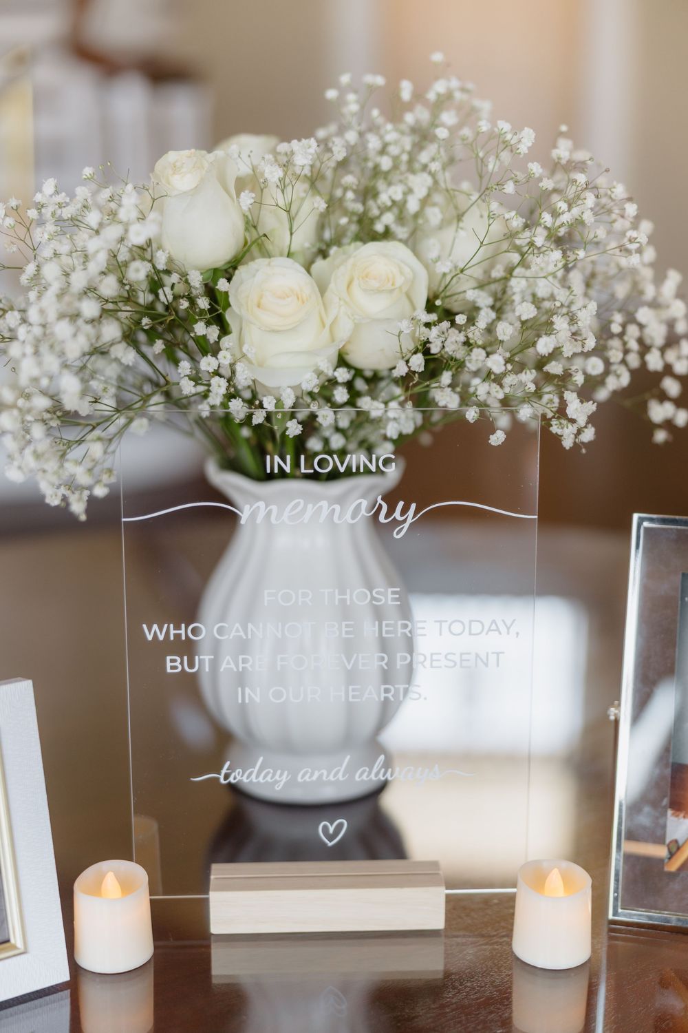 Elegant wedding memorial table with white roses, baby's breath, candles, and an 'In Loving Memory' acrylic sign.
