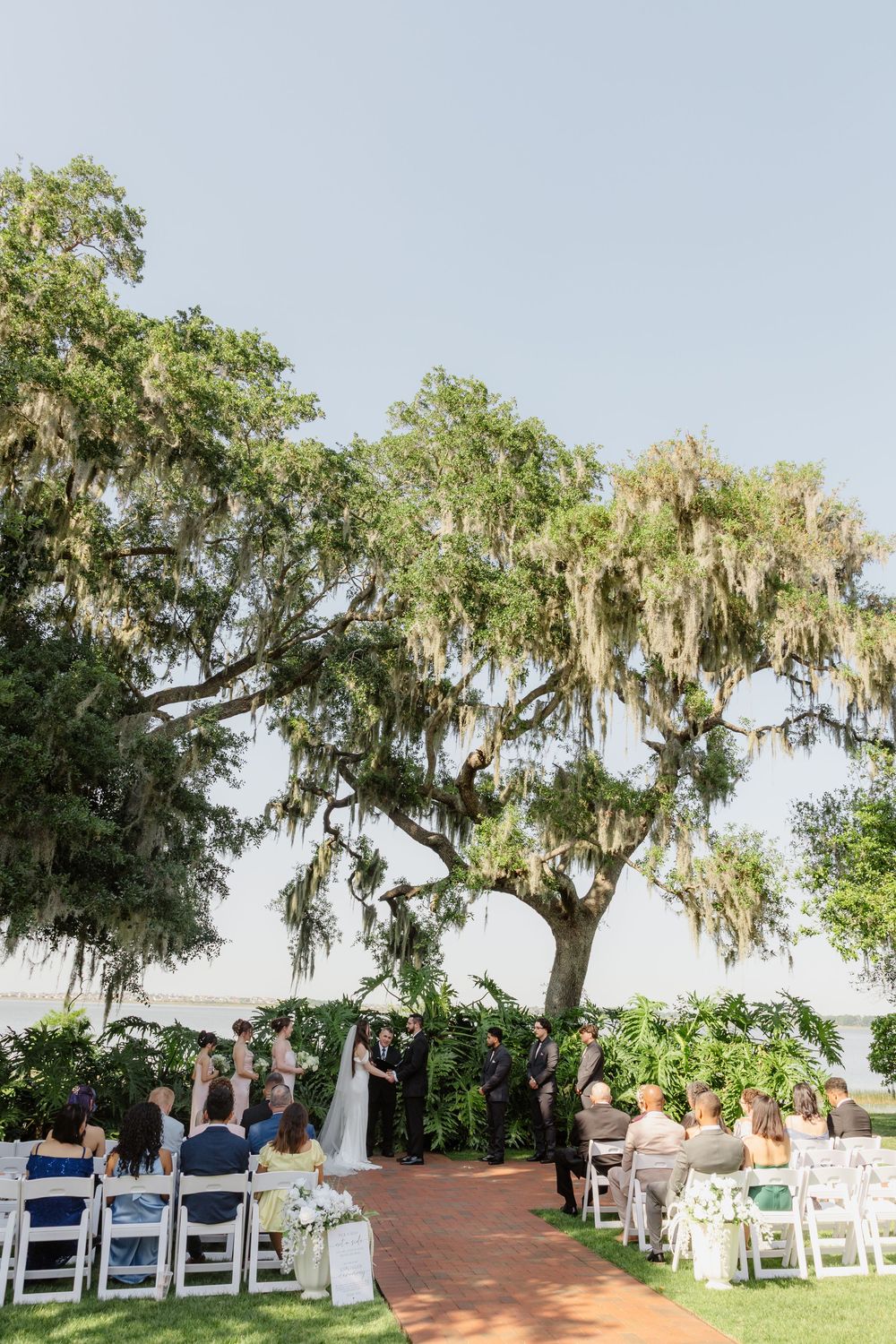 Guests seated in white chairs witness an outdoor wedding ceremony beneath a grand Spanish moss-covered oak tree.