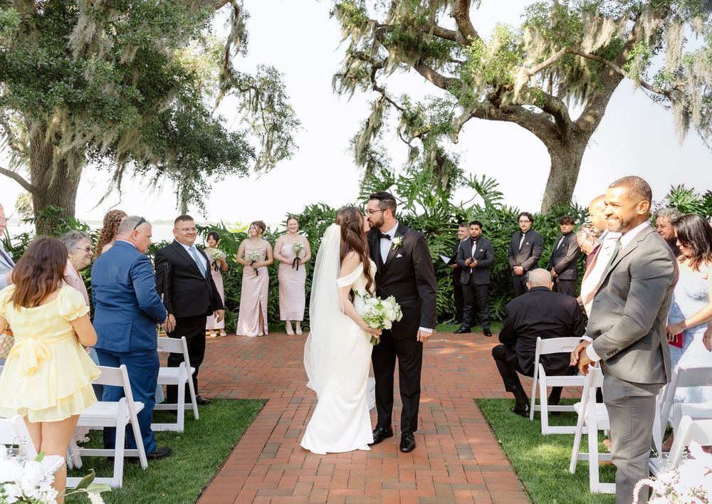 Bride and groom share a kiss at outdoor wedding ceremony on brick aisle lined with white chairs and guests.