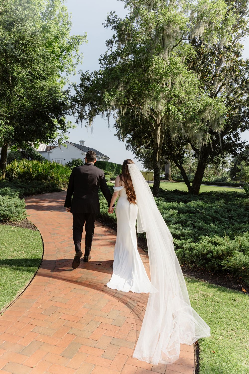 Bride and groom walk hand-in-hand along a brick path surrounded by lush green trees and gardens.