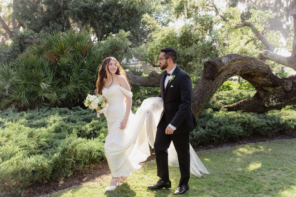 Bride and groom walking through lush garden, bride in off-shoulder gown holding white bouquet, groom in black tuxedo.